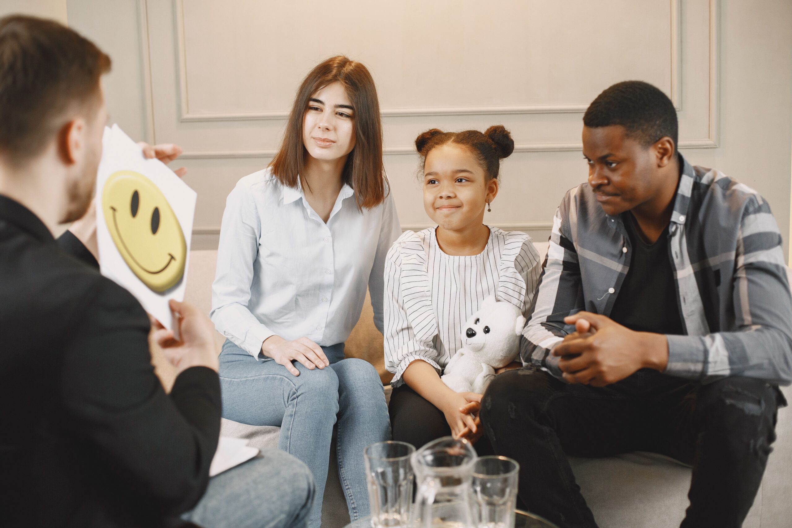 Family therapy session — therapist holds a smiley face card while parents sit with their young daughter holding a teddy bear